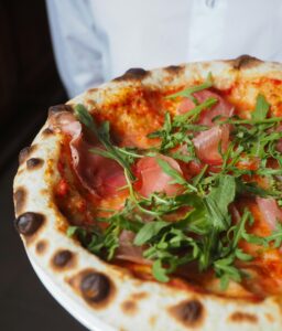 Parmigiana Pizza – a delicious Italian pizza topped with tomato, fior di latte mozzarella, Parma ham, fresh rocket, and flaked Parmigiano Reggiano, a flavorful choice from Sicily Restaurant’s menu.